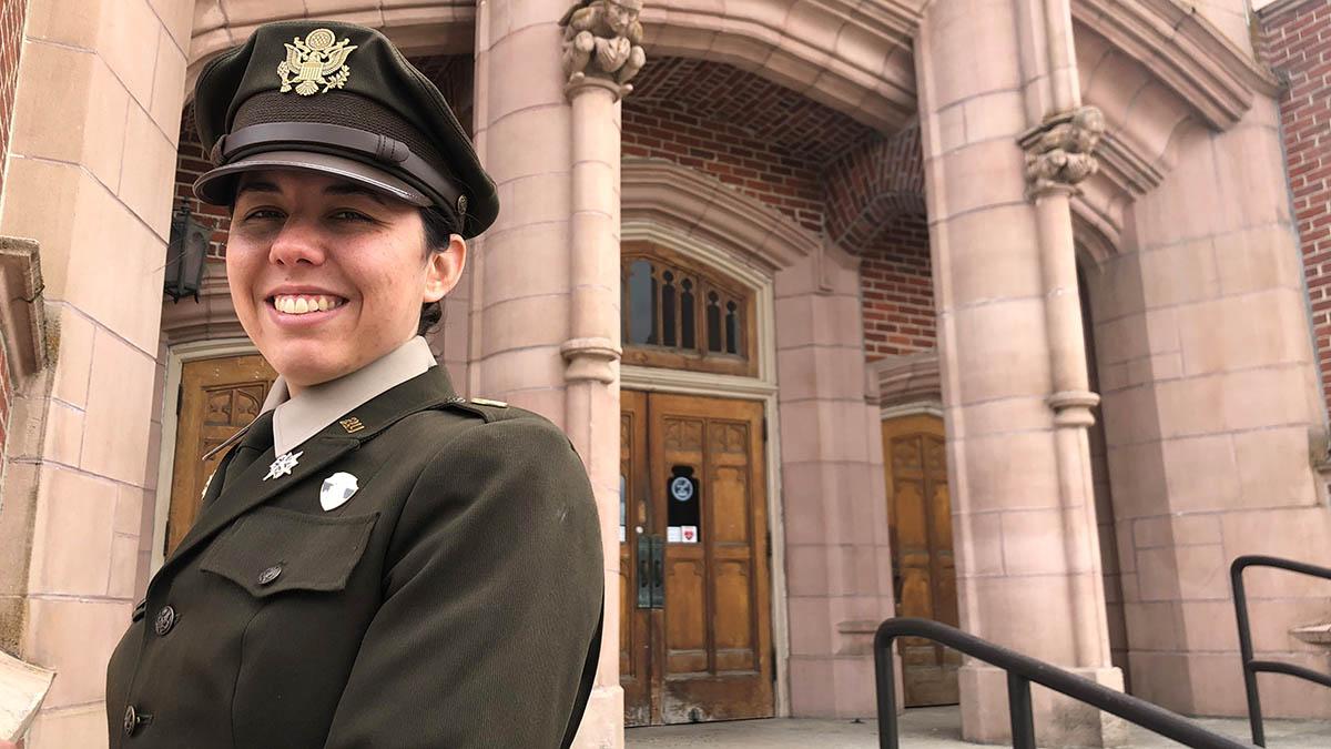 Woman in Army green dress uniform stands in front of brick building.